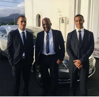 Trois agents funèbres élégants en costume devant un corbillard gris.